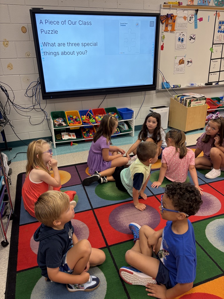 A group of children sit on a colorful floor mat in a classroom, attentively watching a large screen.
