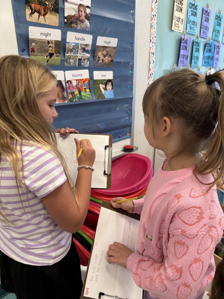Two young girls are indoors, one holds a clipboard and pencil, the other holds paper. They are both focused on their activities. Behind them is a bulletin board with pictures and words.