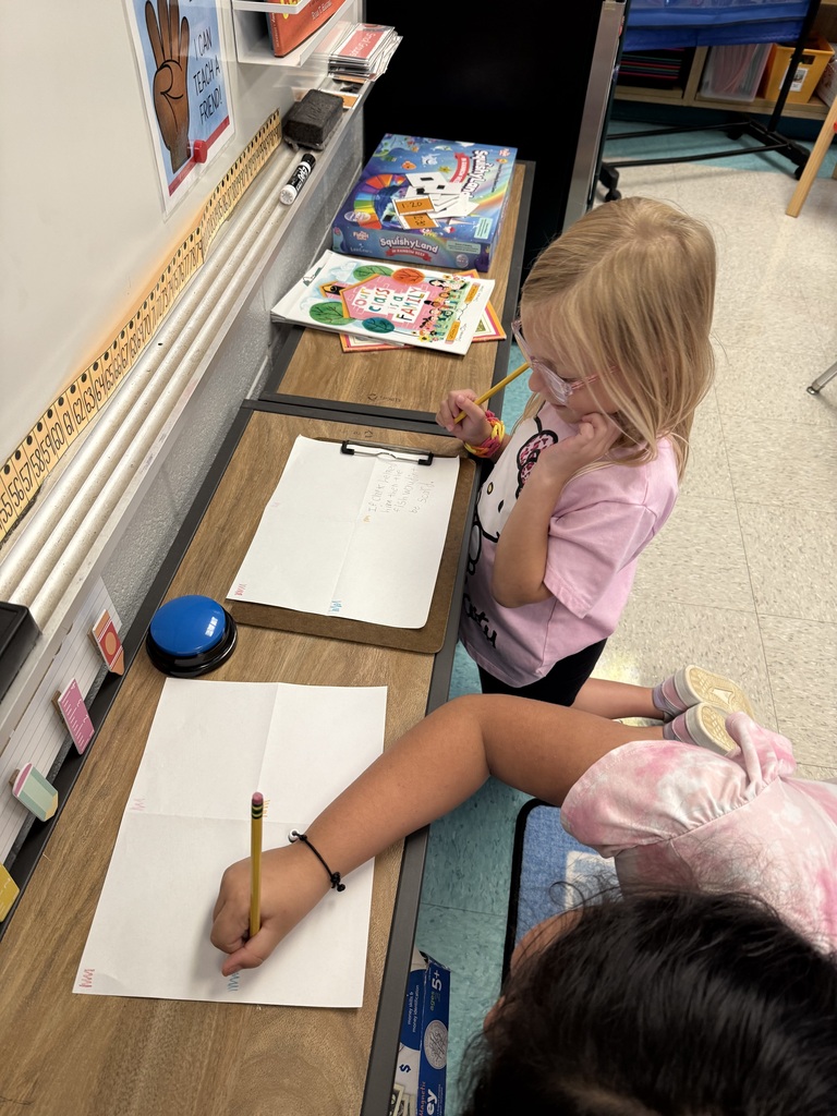 Two young children at a desk writing on paper with pencils. One child has glasses. Background contains a whiteboard and books.