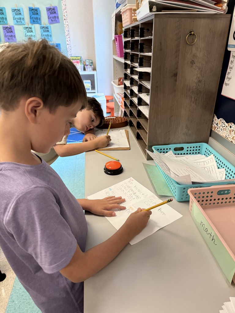 Two children write in a classroom with a shelf and paper baskets. One child uses a stamp.