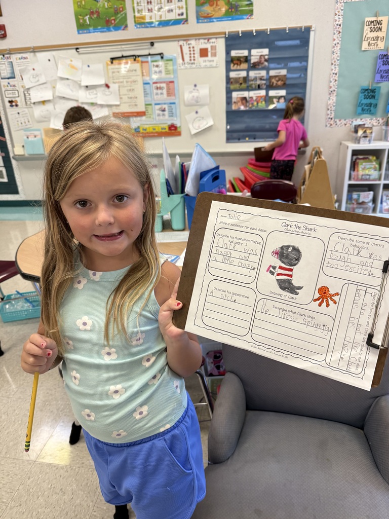 Young girl in a classroom holding a clipboard with a drawing of an animal and text.