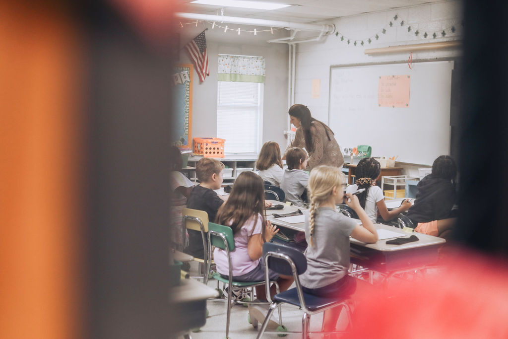 Students in chairs facing desks, a teacher walks by. Bright room with a whiteboard and flag.