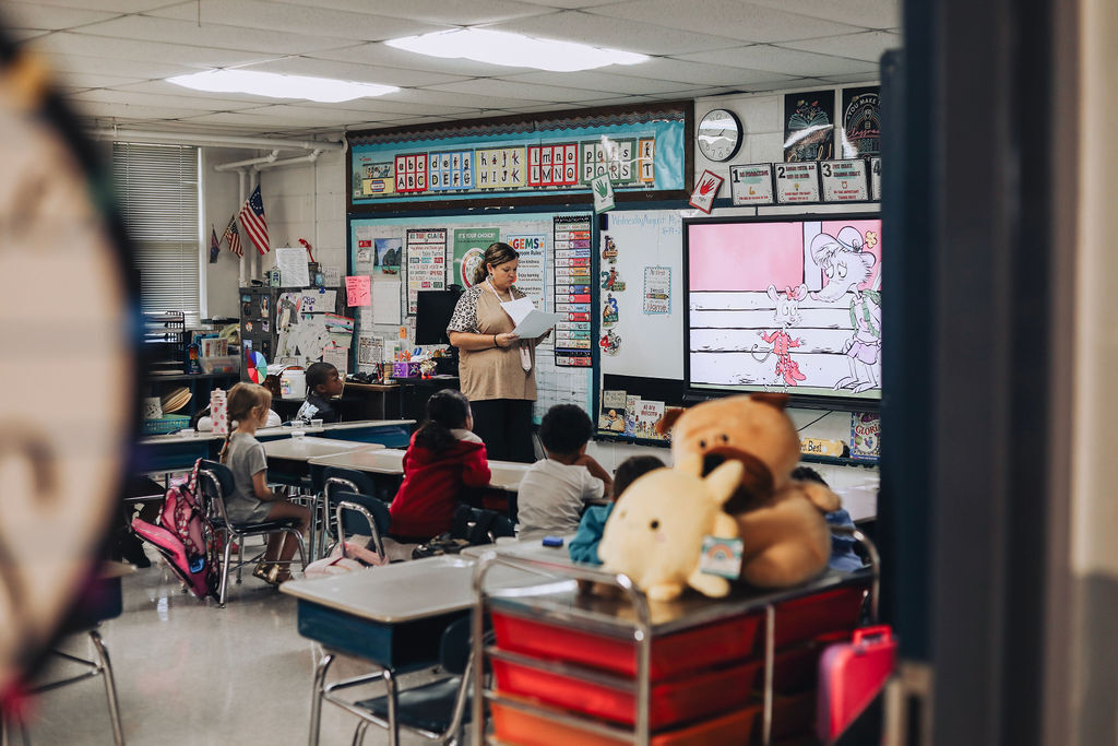 A classroom with a teacher standing by a projector screen. Students sit at desks with stuffed animals and school supplies around them.