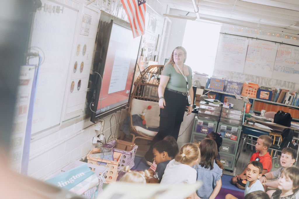 A woman stands in front of a classroom with children seated. The classroom has shelves, a flag, and a whiteboard.