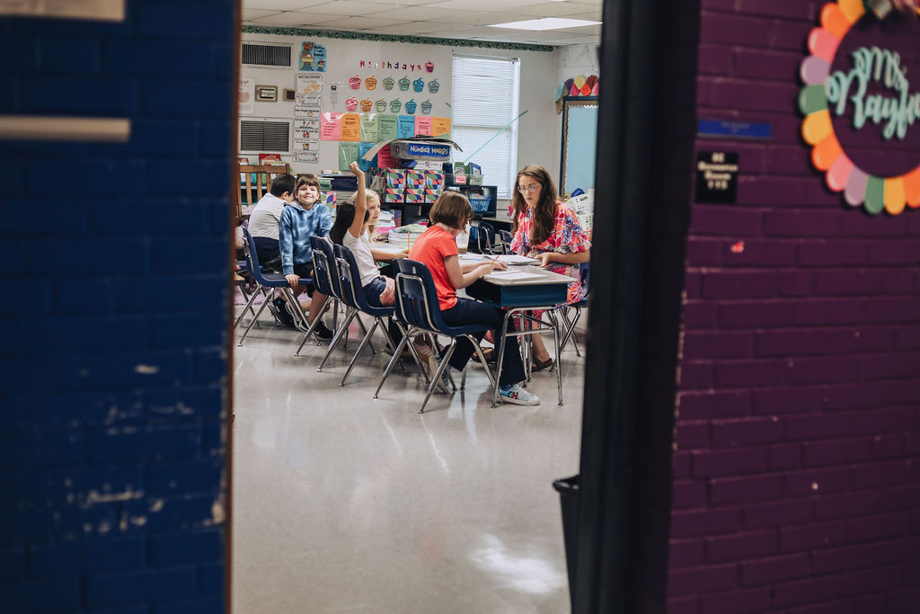 Classroom with children seated at tables. A teacher stands nearby. Colorful decorations on walls. Bright, open space.