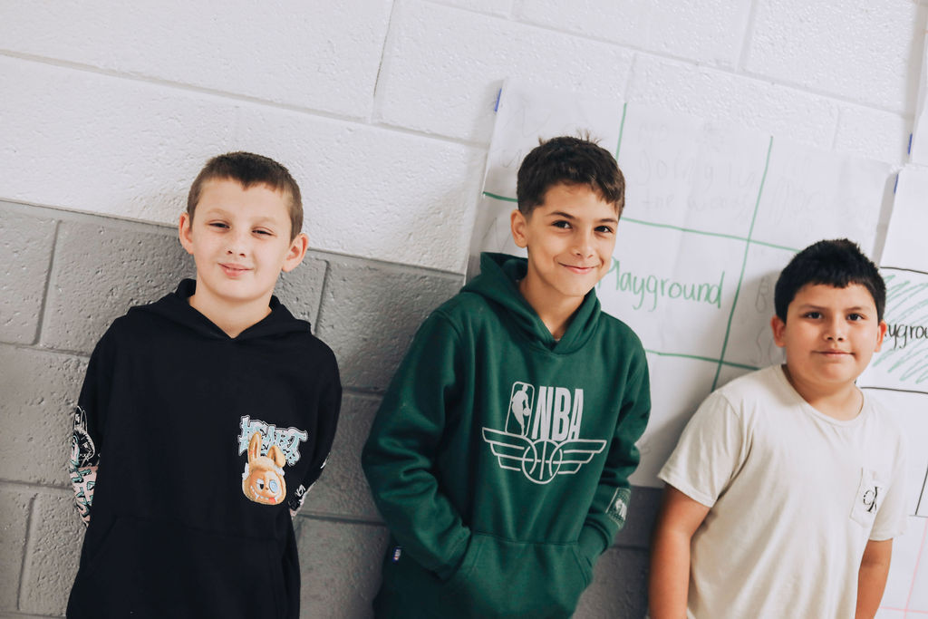 Three boys stand together against a white wall, one in a black hoodie, one in a green hoodie, and one in a white shirt.