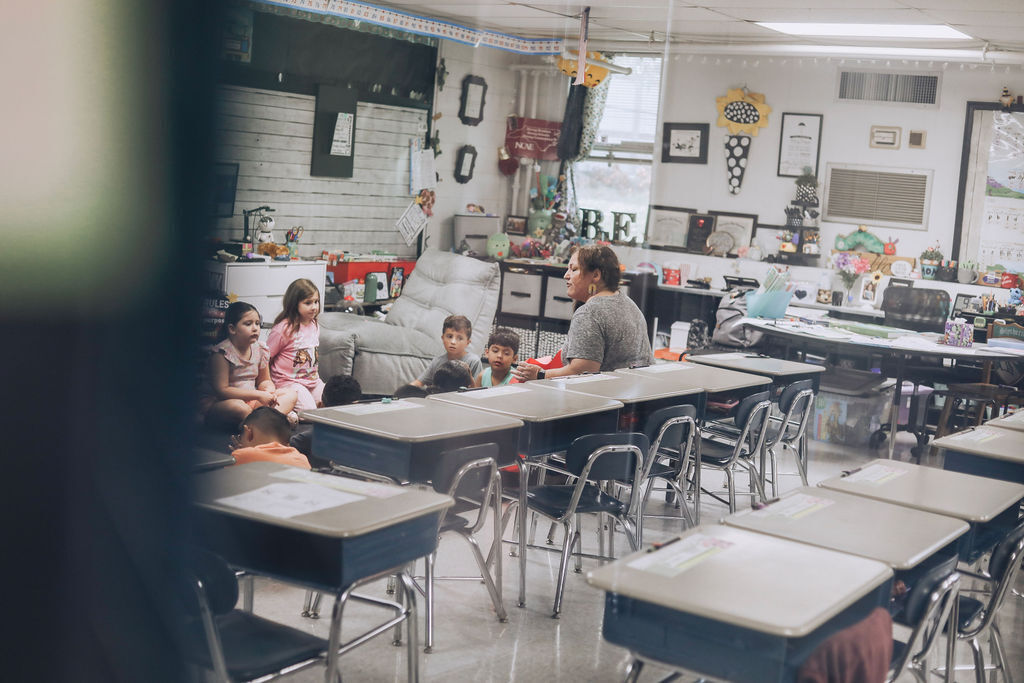 A classroom with a teacher seated at a desk. Students are sitting at desks around the classroom.
