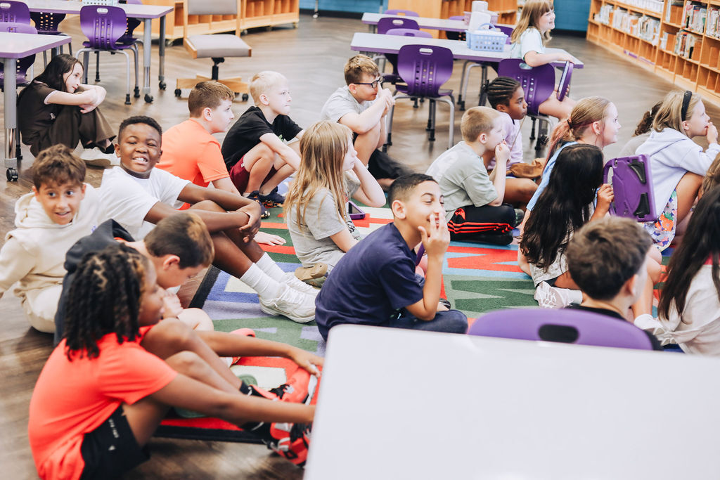 Group of children sitting on a colorful rug in a library. Some sit on chairs, while others are on the floor.