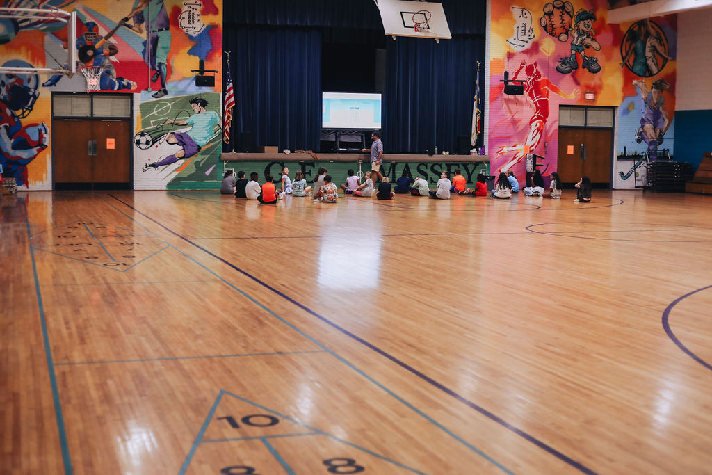 Gymnasium with colorful murals. Kids sit in a circle in front of a person. Blue lines on the floor.