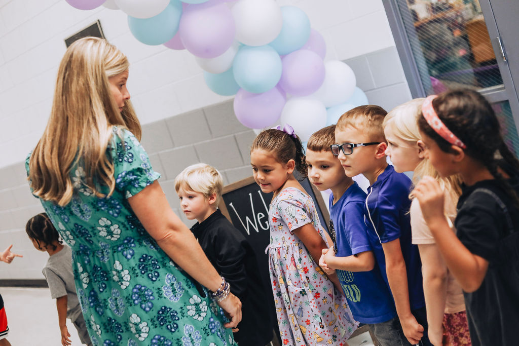 A group of children and an adult woman stand together in a room with balloons in the background.