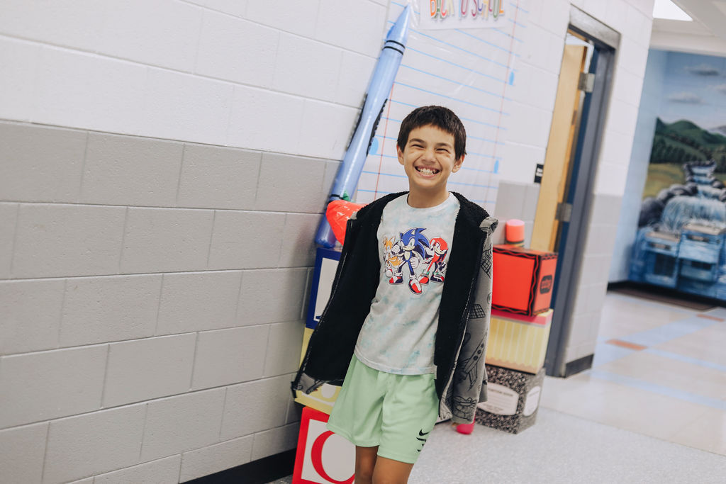 A young boy in green shorts and a jacket smiles in a hallway with a rocket prop.