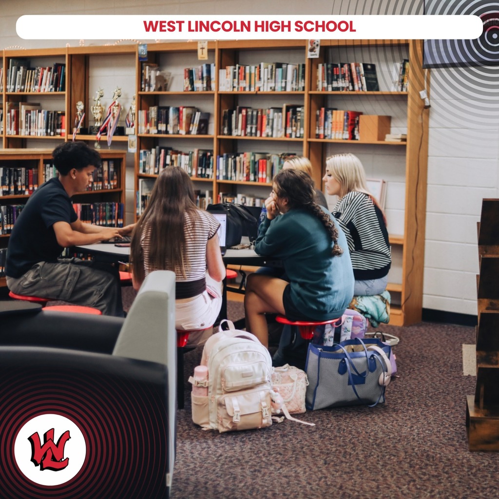 Five students sit around a table in a library, with backpacks and bags on the floor.