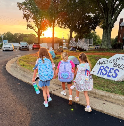 Three children, each carrying backpacks, walk on a sidewalk at sunrise. A large rock with text reads "WELCOME BACK RSES."