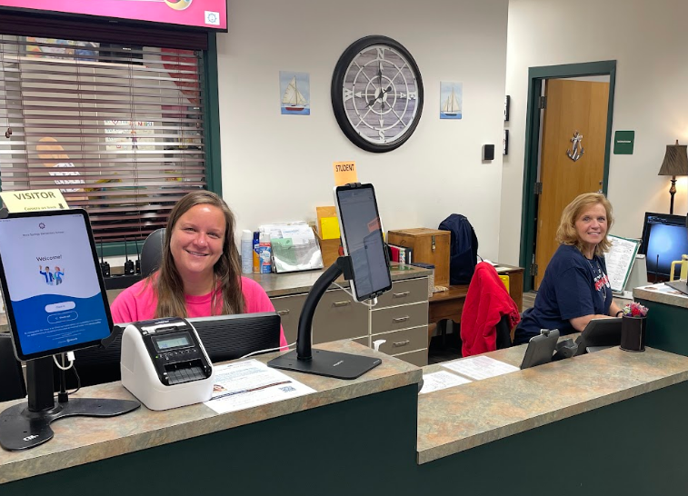 Two women sit at a reception desk, one smiling, with monitors and a clock on a wall behind them.