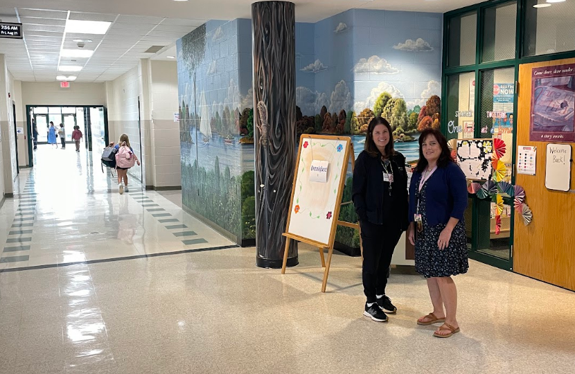 Two women stand next to an easel in a school hallway, with mural backgrounds and bulletin boards.