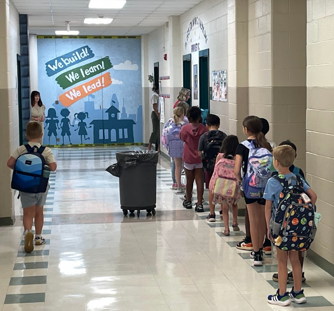 Hallway with a mural. Students line up with backpacks. A trash bin sits in the middle.