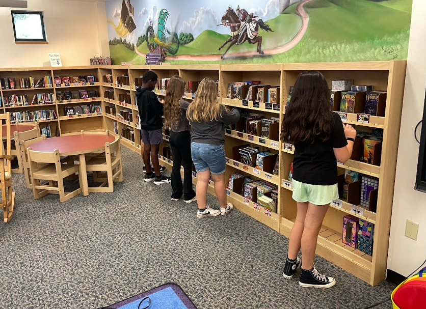 Four children stand in a library. They are looking at books on shelves. A mural is behind them.