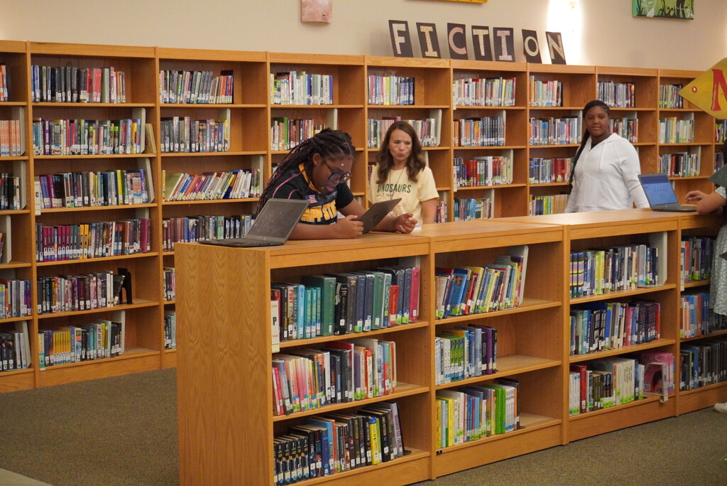 Three people in a library, one seated using a laptop and two standing near a counter with books on shelves.