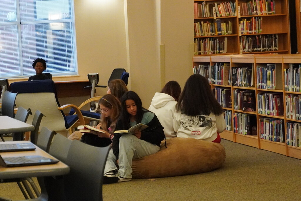 A group of people reading books in a library. Bookshelves and tables are in the background.