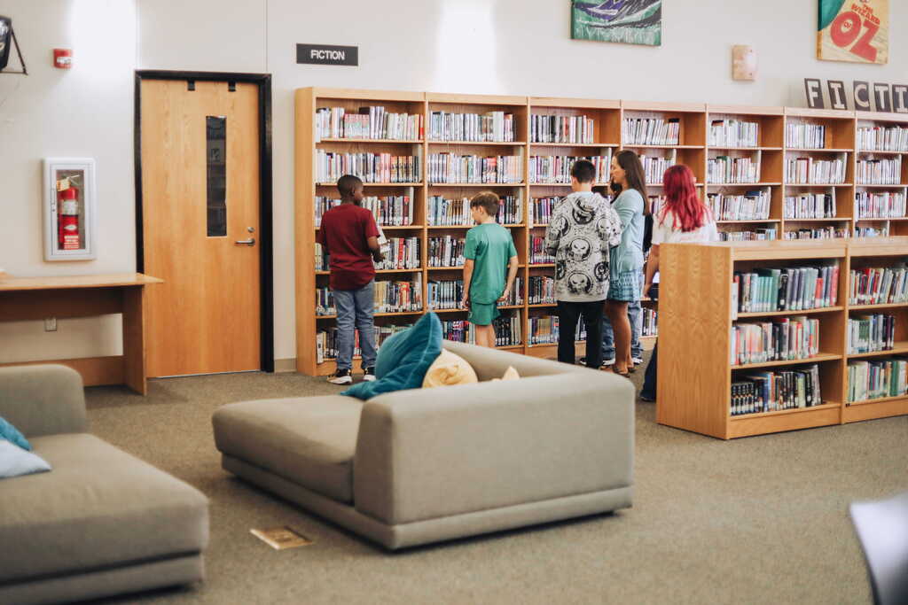 A library with shelves of books. Several people, including children, are standing and looking at the books.
