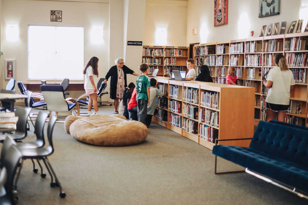 A library interior with people standing and sitting. Shelves filled with books. A bean bag chair on the floor. A blue velvet sofa.