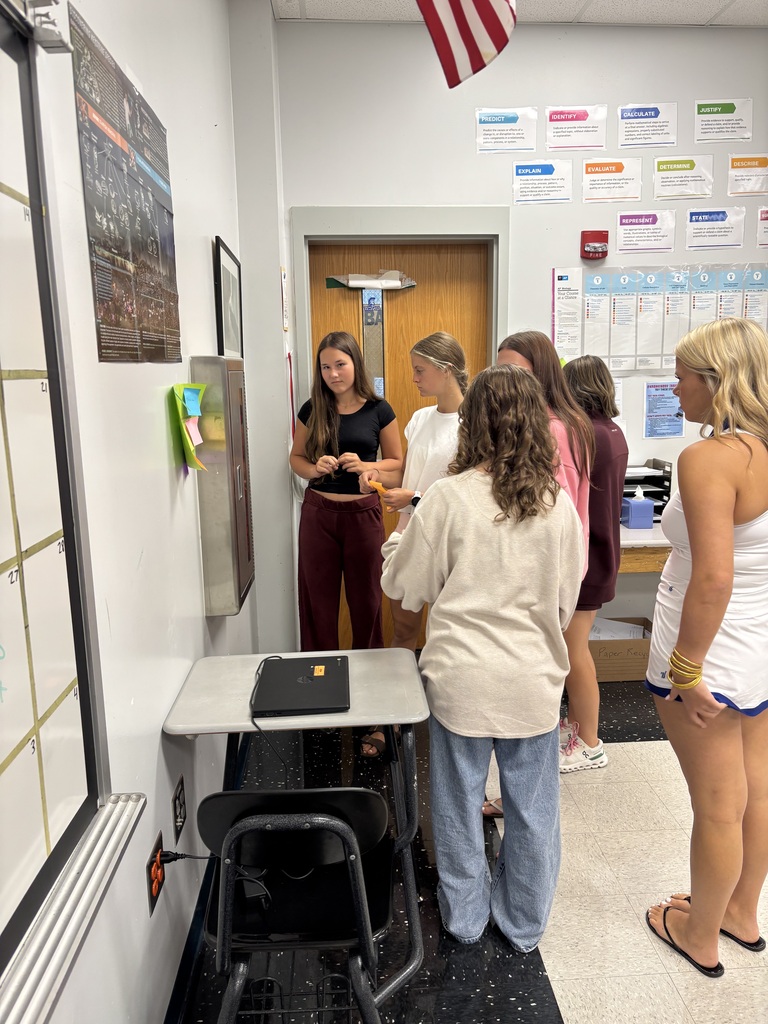 Six people stand in a room. A woman talks to a person in a black shirt. A table holds a laptop.