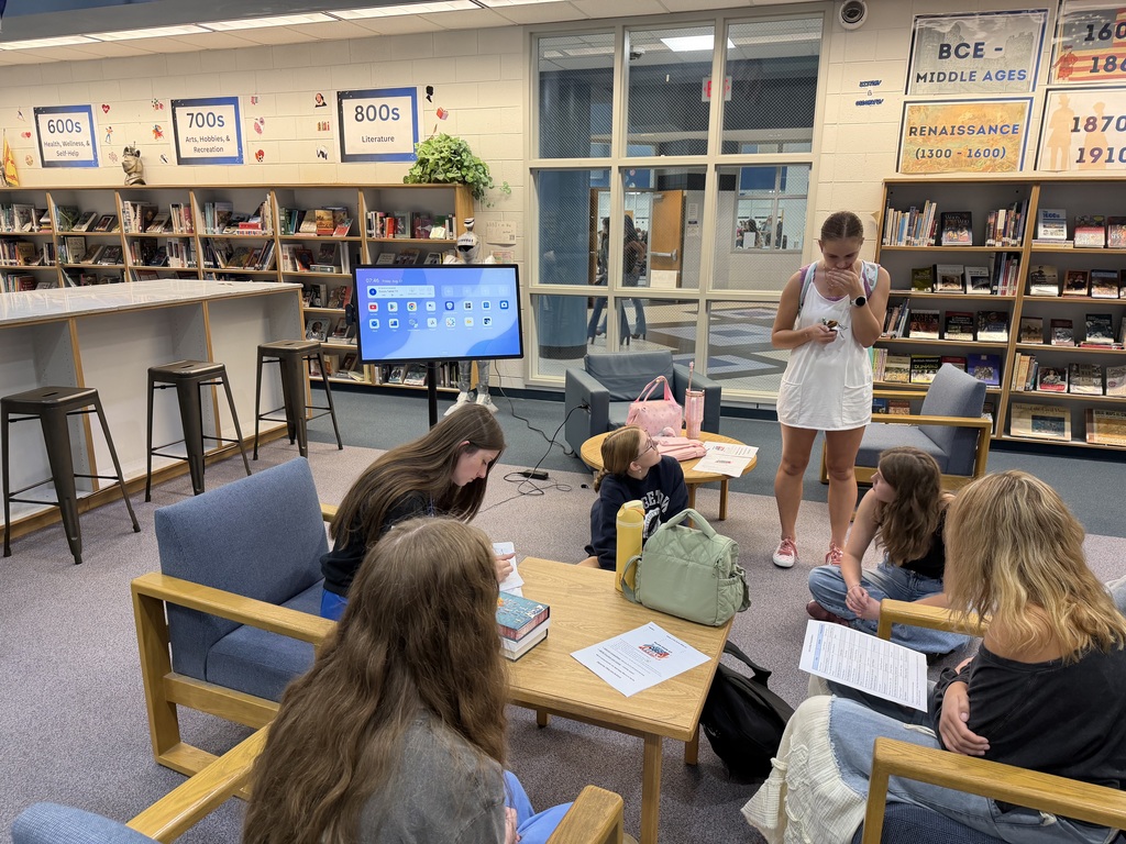 Library reading group with people seated at tables. Woman stands in front of a screen. Shelves with books in background.