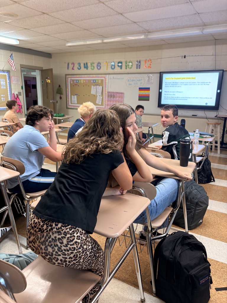 Students sit at desks in a classroom, facing each other in groups. A television is mounted on the wall.