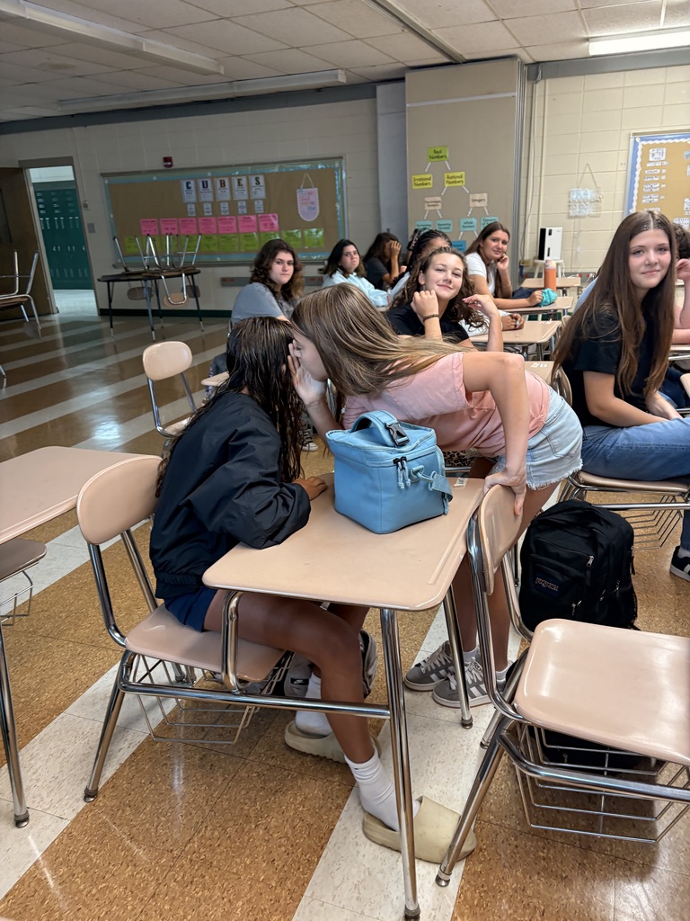 A classroom with desks; students seated, one leans over a desk while another sits behind her. Backpacks and bags are on the floor.