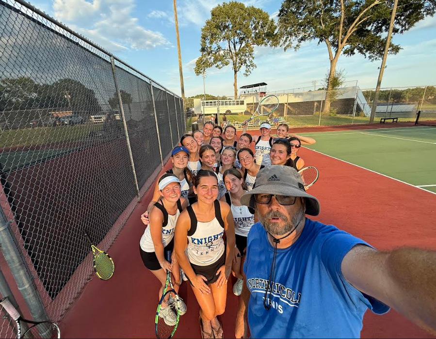 A tennis coach and his team pose for a group photo on a tennis court.