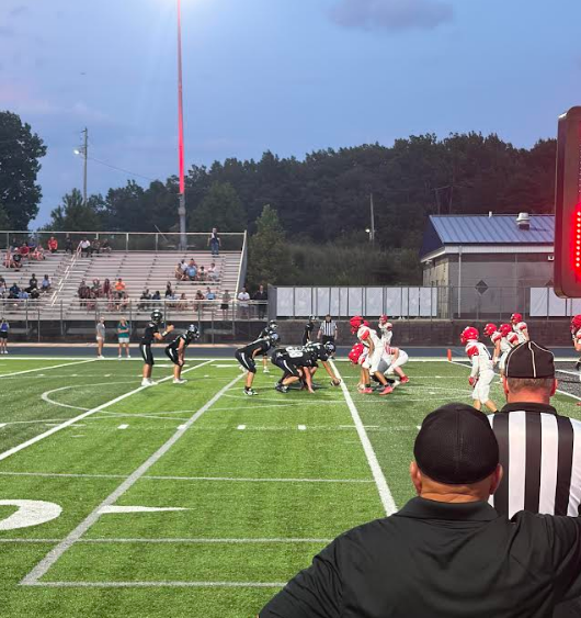 Football game in progress, players on field, spectators in stands, score board visible.