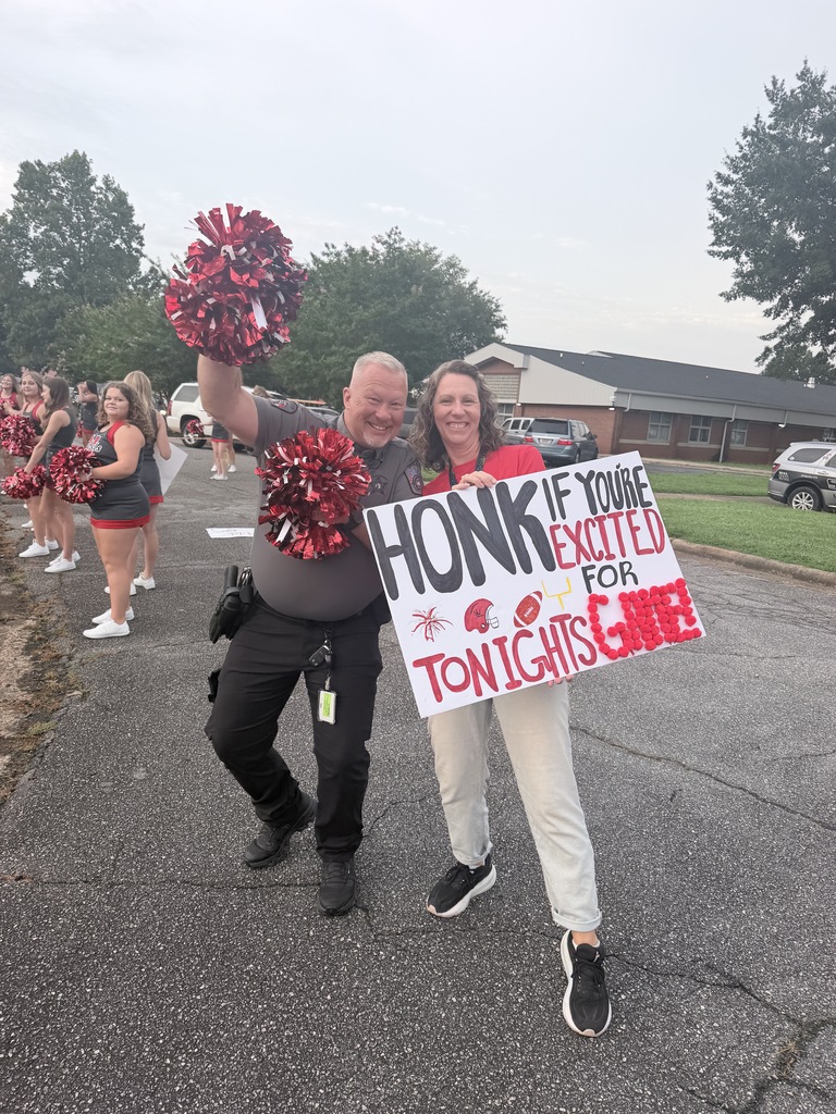 Two people stand in a street, holding a sign and pom-poms. A woman wears a red shirt, and a man holds red pom-poms. A group of cheerleaders and buildings are in the background.