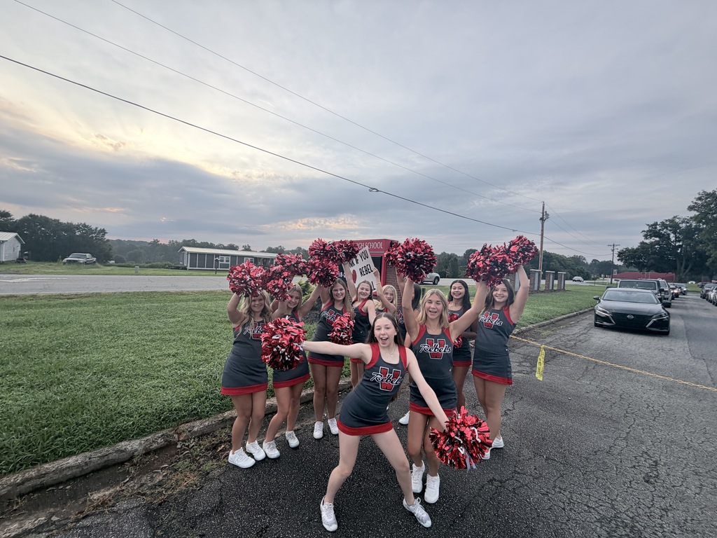 A group of cheerleaders in red and black uniforms holding pom-poms in a road setting, with a car parked nearby.