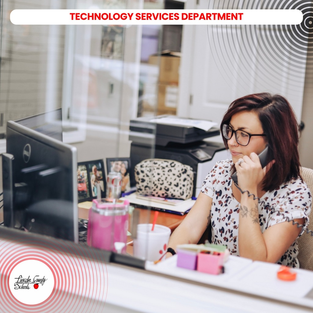Woman sitting at a desk, talking on a phone. Behind her is a glass partition, a printer, and office supplies.