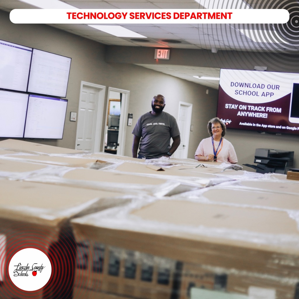 Two people stand behind boxes. A sign reads "Download Our School App". A monitor is on the wall.