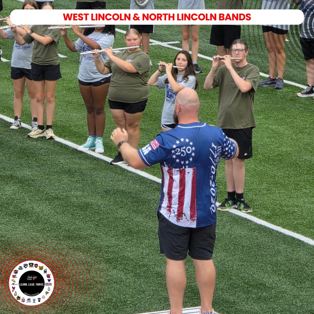 Several people stand on a field, holding instruments, while a man in a blue shirt leads them.