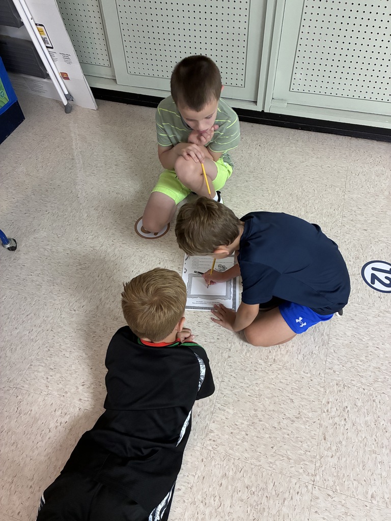 Three boys work on a project on the floor, one sitting, two kneeling. One holds a paper, another holds a pencil.
