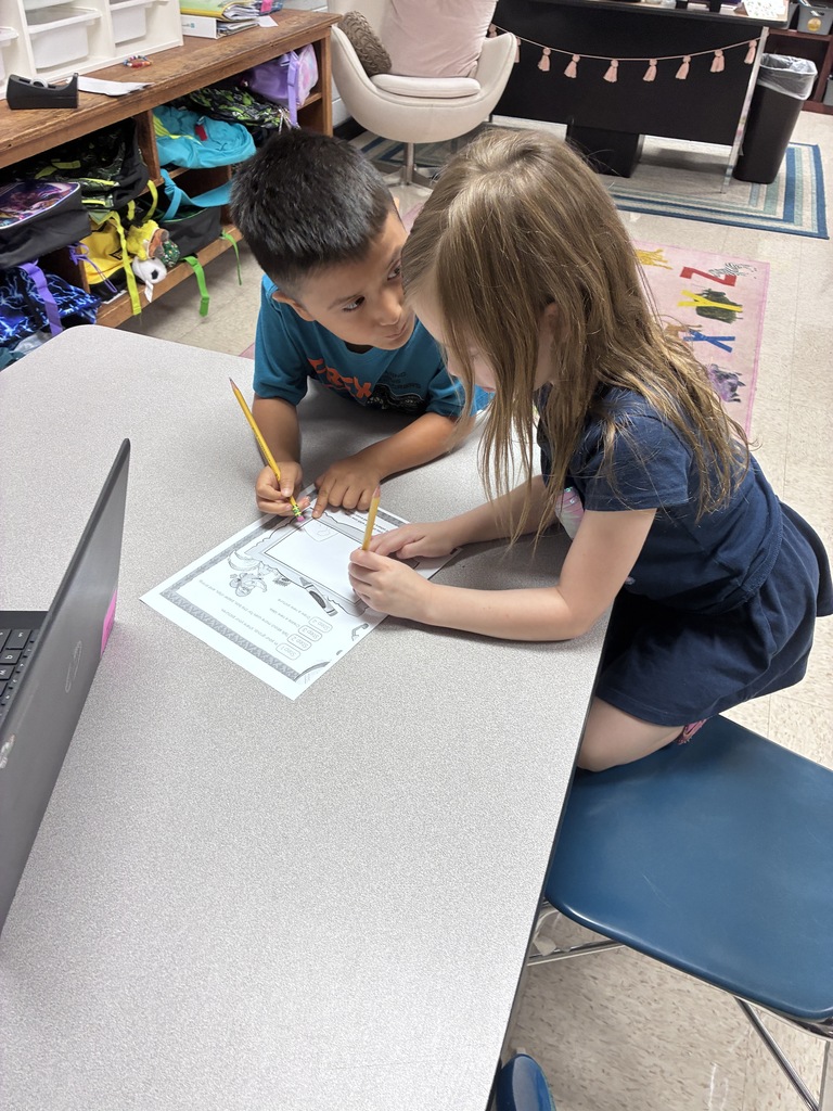 A boy and a girl sit at a table, focused on a worksheet with pencils. A laptop rests on the table, and chairs are nearby.