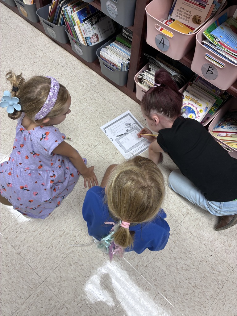 Three children in a classroom, two sitting on the floor with a paper, one holding a pencil. Shelves filled with books behind them.