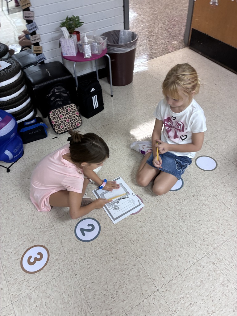 Two children sit on floor, one holding book, other writing on paper. Behind, items like a trash can and table.