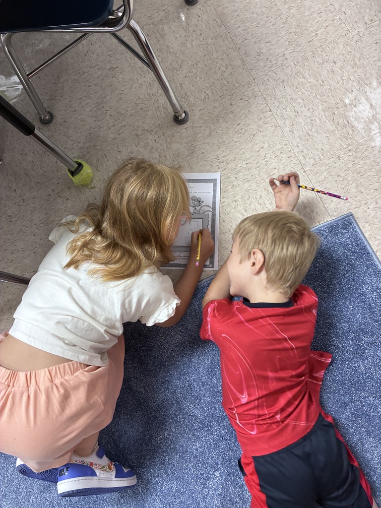 Two children are sitting on a blue mat, one using a pencil and the other using a crayon.
