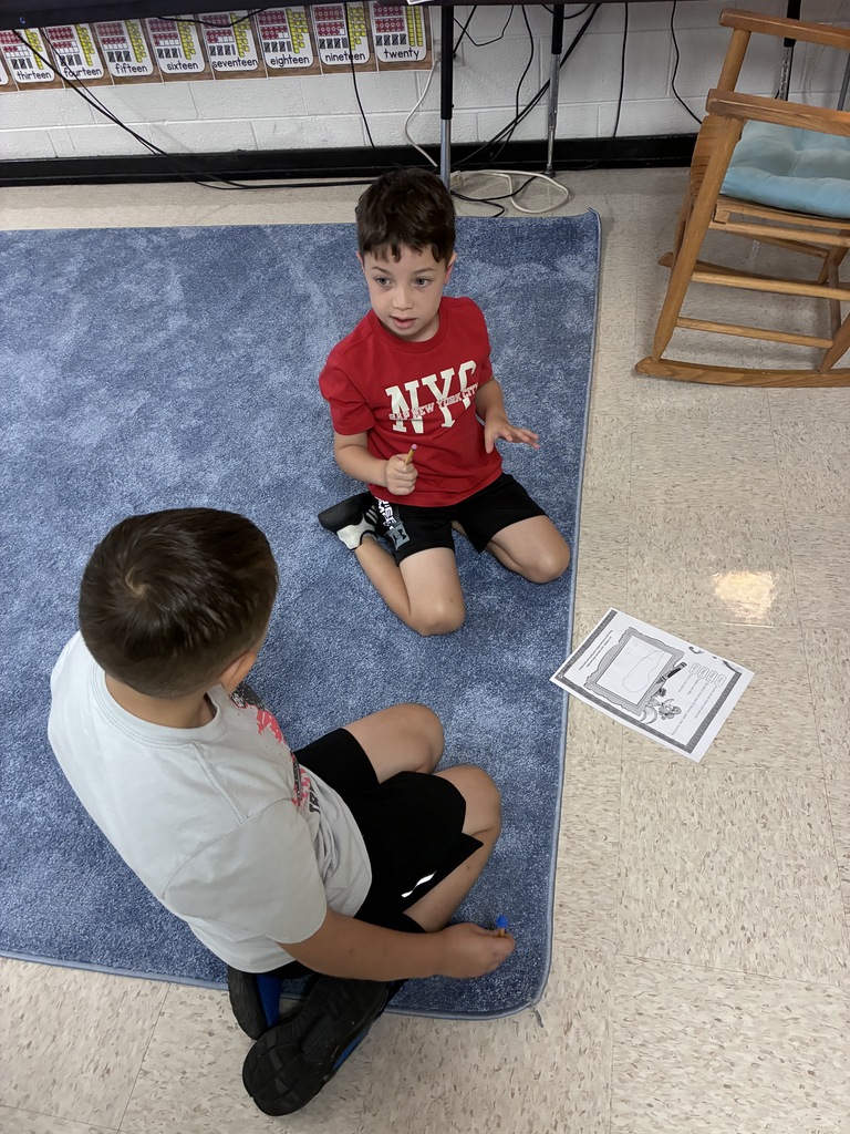 Two boys in a classroom sit on a blue carpet. One boy is gesturing with his hands.
