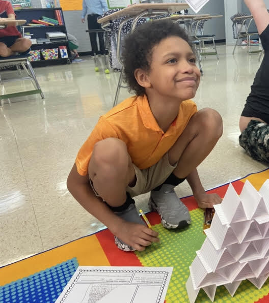 A child kneels on a colorful floor mat, constructing a pyramid from white papers. Behind, other students are present in a classroom setting.