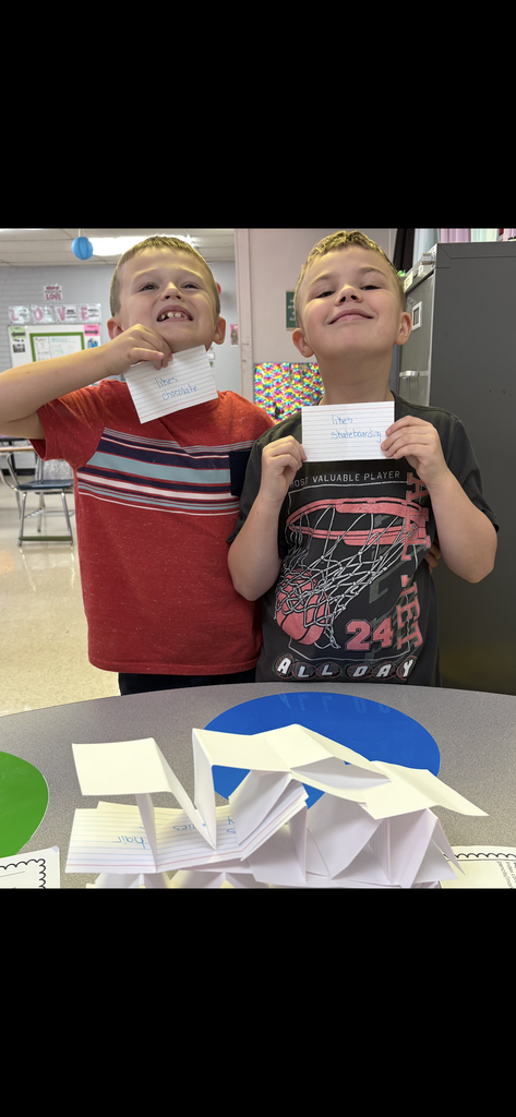 Two children, one in a red shirt and one in a black shirt, smile while holding papers in a classroom.