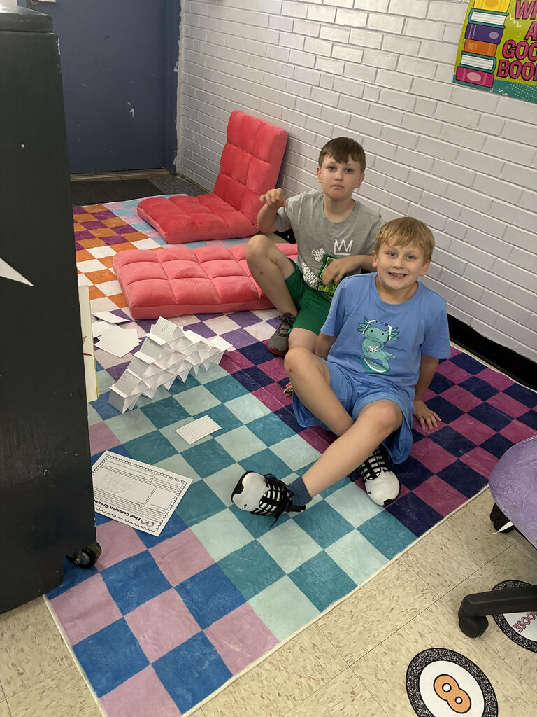 Two boys sitting on floor with a colorful rug, one holding a paper, and another wearing a grey shirt.