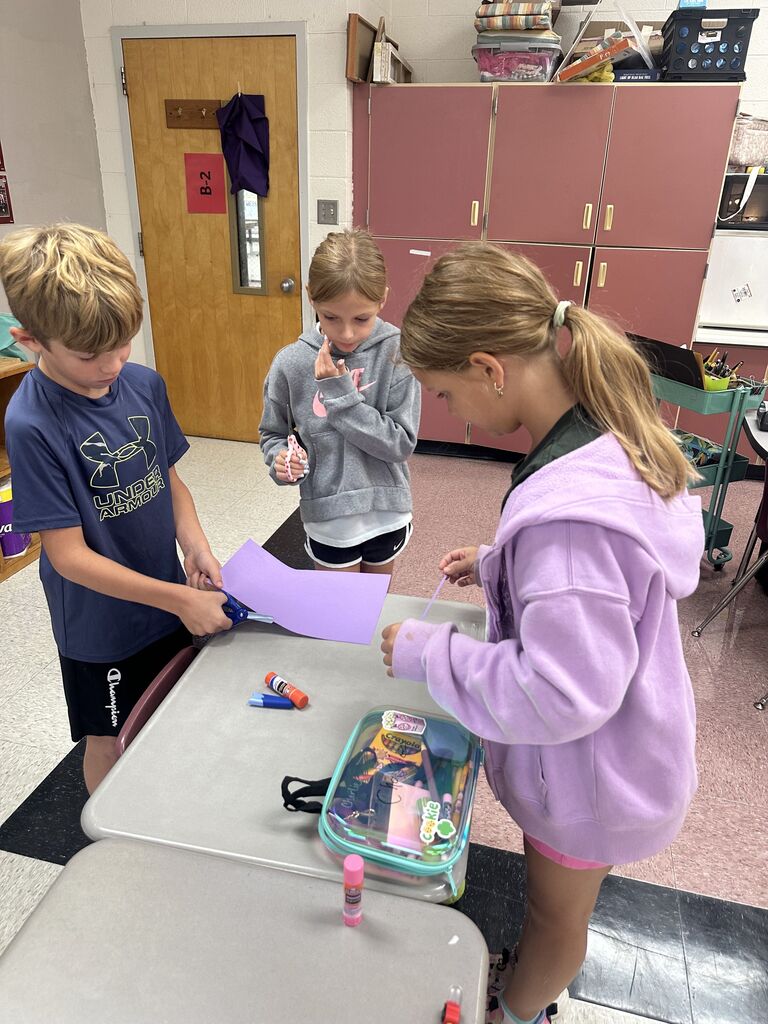 Three elementary students working together around a classroom desk on a hands-on project. A student in a blue Under Armour shirt cuts purple construction paper, a student in a grey Nike hoodie holds scissors while observing, and a student in a light purple zip-up hoodie works with paper strips near a clear pencil case.