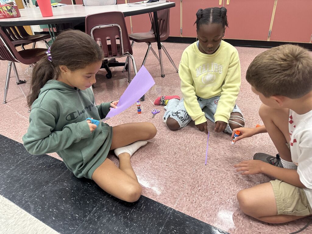 Three elementary students sitting cross-legged on a tiled classroom floor working collaboratively on a craft project. A student on the left in a green hoodie uses scissors to cut purple construction paper, while a student in a yellow sweater and a student on the right focus on gluing paper strips to the floor.
