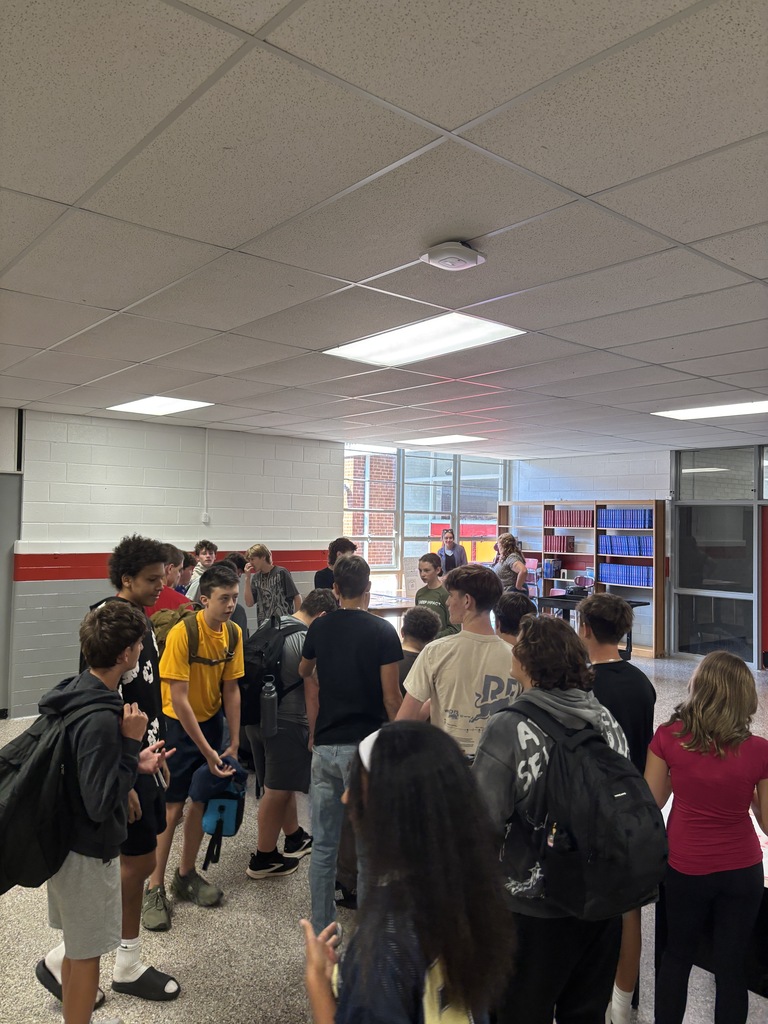 A hallway with people standing around, some carrying backpacks. Ceiling has recessed lights, and a door is in the background.