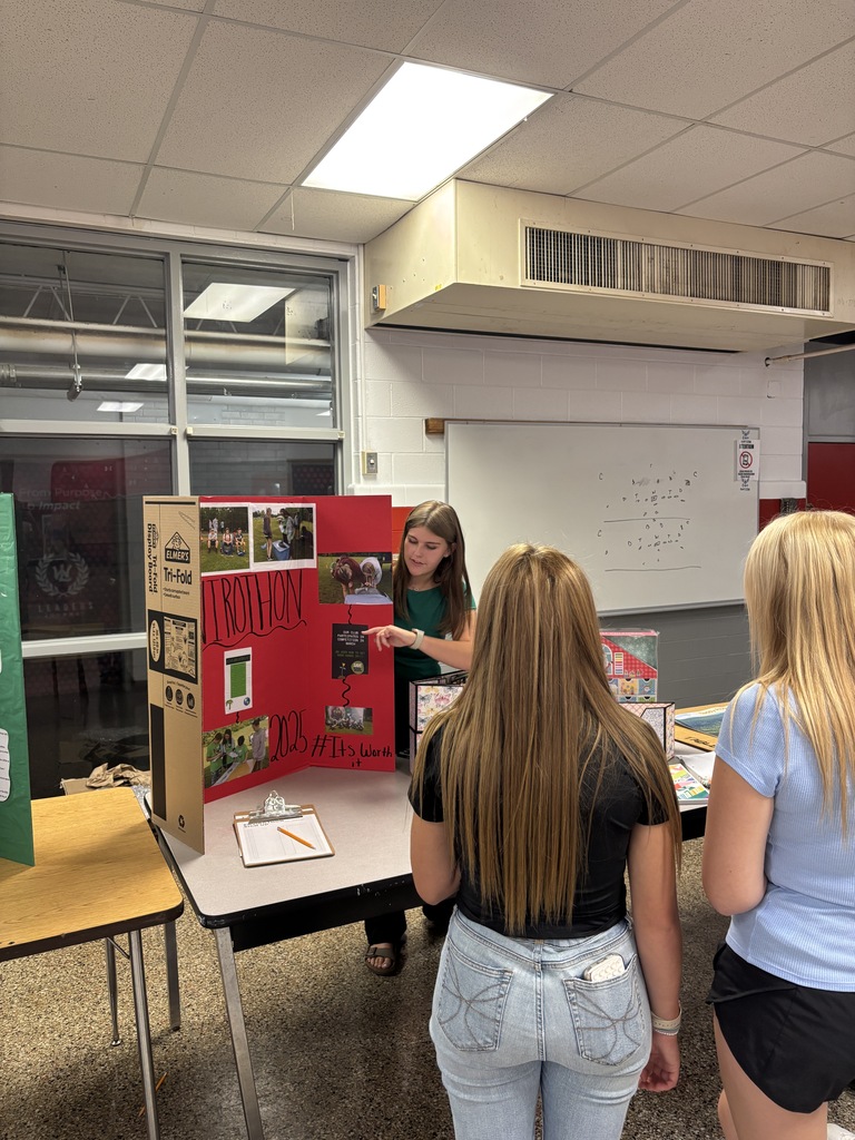 Three women stand around a table displaying a poster, with one woman in green holding a pen.