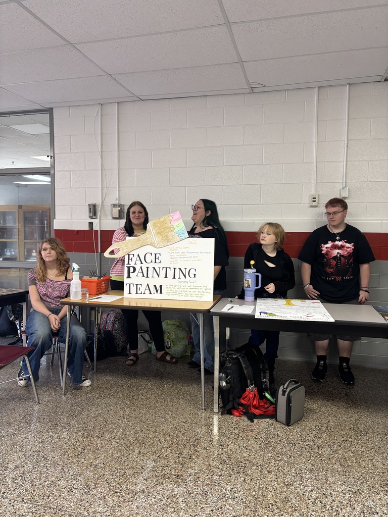 A group of people are gathered around tables in a room. One table has a sign that reads "FACE PAINTING TEAM".